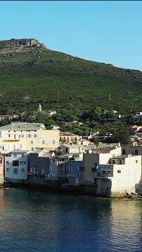 Charming waterfront houses line the coast of Erbalunga, Corsica, with lush green hills rising in the background under a clear sky.