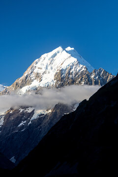 View of a snow-capped mountain peak piercing the azure sky, its slopes touched by soft clouds and dark shadows, creating a striking contrast, Mount Cook, Canterbury Region, New Zealand.