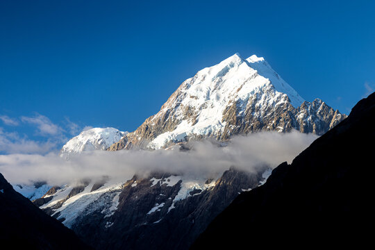 View of a majestic snow-capped mountain peak piercing the clear blue sky, partially veiled by ethereal clouds, Aoraki Mount Cook, Canterbury Region, New Zealand.