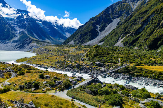 View of a glacial river winding through a golden valley towards snow-capped mountains under a vibrant blue sky, framed by rugged terrain, Mount Cook, Canterbury Region, New Zealand.