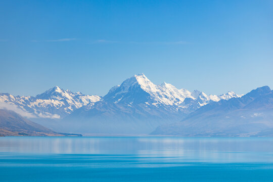 View of snow-capped mountains mirrored on the tranquil, turquoise lake, under a clear blue sky, a serene landscape evoking peace and adventure, Lake Pukaki, Mount Cook Lookout, New Zealand.
