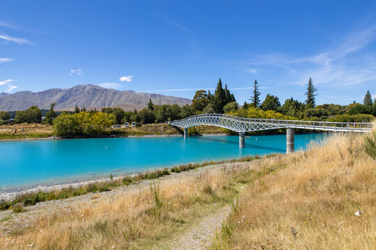 View of vibrant turquoise waters flow beneath a white bridge under a clear blue sky, framed by golden grasses and distant mountains, Lake Tekapo, New Zealand.