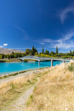 View of a brilliant turquoise river flowing beneath a sleek modern bridge, contrasting with the golden grasses under a clear blue sky, Lake Tekapo, New Zealand.