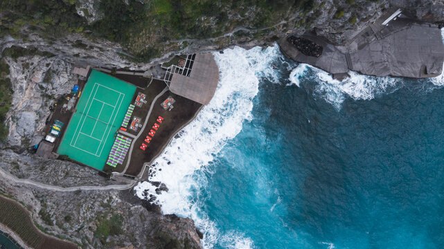 Aerial view of a vibrant green tennis court perched atop rugged cliffs, where crashing turquoise waves meet the rocky shore, Laurito, Campania, Italy.