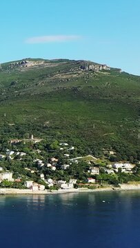 Aerial view of a picturesque coastal village nestled between lush green hills and the calm blue sea in Erbalunga, Corsica. Bright, clear sky enhances the Mediterranean landscape.