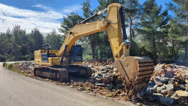 A large yellow excavator is stationary, with its bucket lowered toward a pile of loose rocks and dirt on a steep embankment next to the road