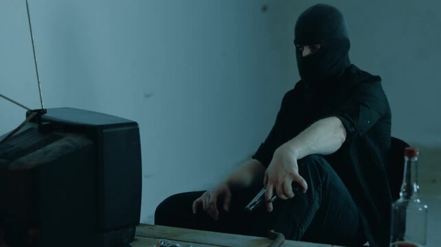 Male kidnapper in a black balaclava sitting at a table in the basement and holding a weapon. Female hostage sitting in front of a man. Kidnaping, crime, violence concept. Slow motion