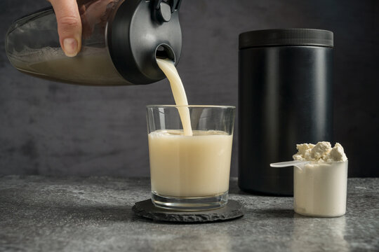 Pouring protein vanilla shake in drinking glass and scoop with whey protein powder on dark background