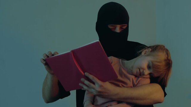 Kidnapper in a black balaclava holding a child in his arms, using book and reading. Little girl sleeping. Copy space. Child abduction concept. Slow motion