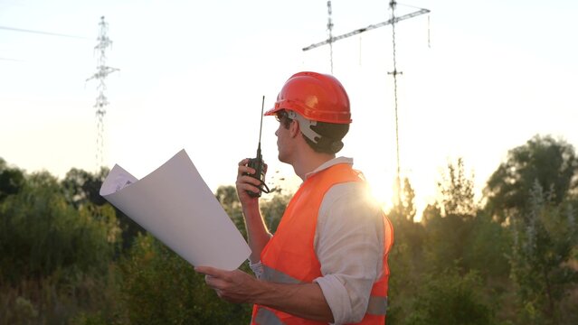 Electrical engineer talking on the walkie-talkie and holding drawing while controlling of repair of electrical high voltage pylons outdoors. Energy industry concept. Slow motion