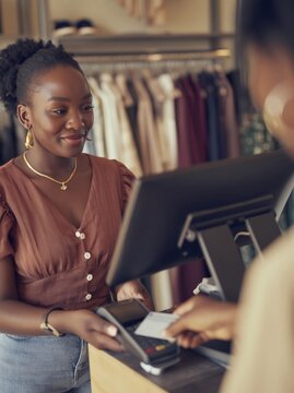 Young South African woman at retail checkout counter, smiling while assisting customer with payment in a clothing store filled with garments and accessories