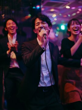 Japanese male singer in black suit passionately performs karaoke with two smiling colleagues in a vibrant, colorful setting filled with lights and reflections