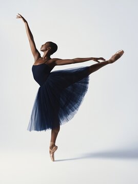Black female ballet dancer in a dramatic pose wearing a dark blue tutu and pointe shoes, showcasing elegance and strength against a clean, light background