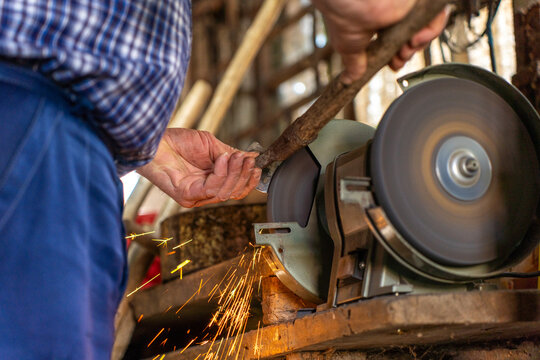 Man sharpening metal tool on electric bench grinder, sparks flying from rotating abrasive wheel in workshop environment.
