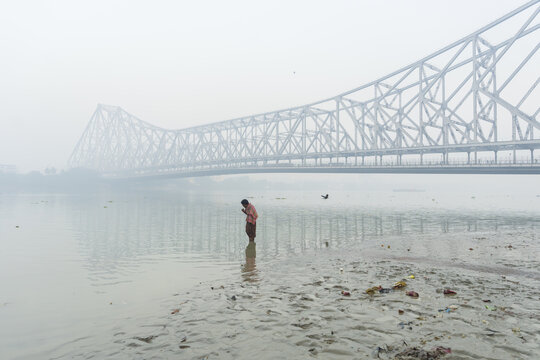 View of a solitary figure stands waist-deep in the tranquil river, their hands clasped in prayer beneath the colossal Howrah Bridge veiled in mist, Kolkata, West Bengal, India.