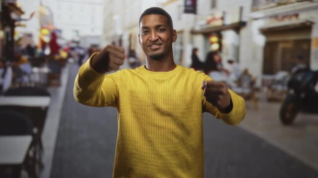 Man in yellow sweater gestures with hands and fists toward camera on a busy street cafe scene; confidence warmth.