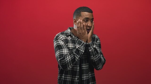 Man with hands on face in studio showing surprised open mouth and wide eyes, wearing plaid shirt against red backdrop; surprise wonder.