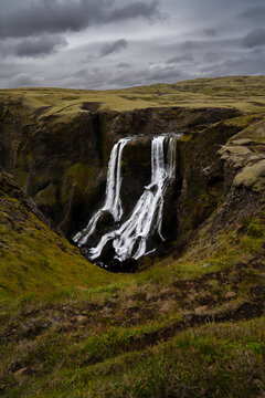 View of water cascades down a rugged cliff face, contrasting with the green vegetation and dramatic sky, creating a mesmerizing landscape, Skaftarhreppur, Iceland.
