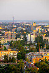 Obraz premium Dnipro, Ukraine, aerial cityscape showcasing a mix of residential buildings, industrial structures like smokestacks and grain silos, with abundant green trees and the river in the hazy distance at ear