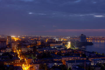 Obraz premium Dnipro, Ukraine, vibrant cityscape at dusk with illuminated bridges spanning the river, showcasing a mix of modern and historical architecture, bustling streets with light trails, and a distant indust