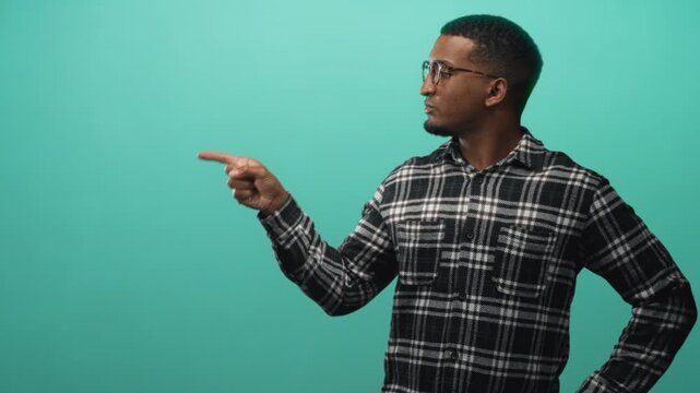 Young black man wearing glasses and plaid shirt points finger and shows open hand palm in studio with teal backdrop; curiosity contemplation.