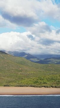 Scenic view of the Delta du Fango coastline in Corsica, featuring sandy beach, lush green hills, and dramatic mountains under a partly cloudy sky.