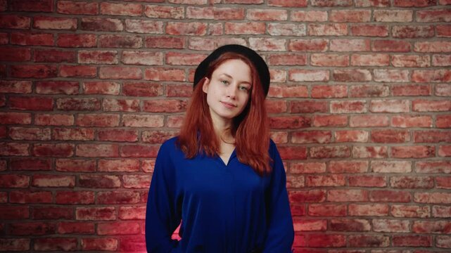 A front view of a pretty young caucasian redhead woman in a blue blouse and black beret, winks and gives a thumbs up gesture against a brick wall. Playful approval and positive reaction