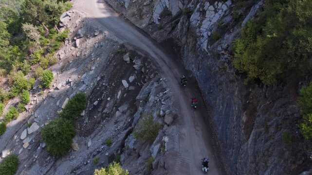 Aerial view of motorcyclists riding on a narrow rocky mountain road with sharp bends and steep cliffs. Adventure touring, remote travel, and rugged nature landscape captured in daylight.