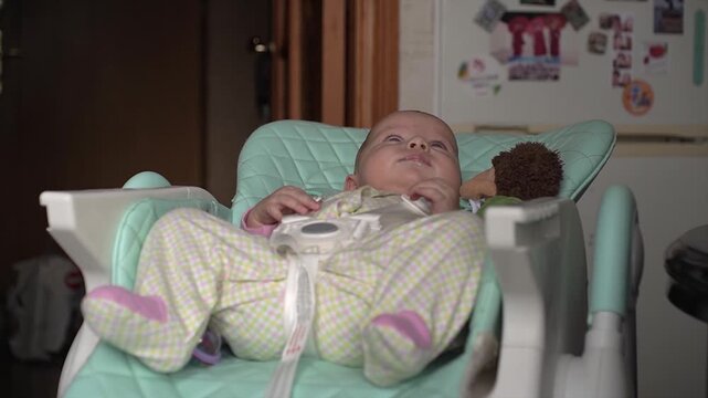 a small, sweet baby, bored in a child seat in the kitchen. He jerks, moves, moves his legs, arms, looks around, shaking with boredom. The camera moves smoothly to the right
