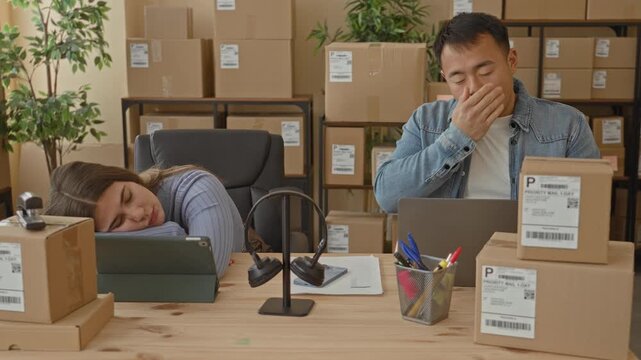Man covering mouth yawning while woman sleeps with head on tablet amid stacked shipping boxes in a packing building; overtime fatigue teamwork.