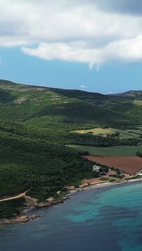 Scenic aerial shot of Plage de Tamarone with turquoise waters, lush green hills, and a dramatic coastline under a partly cloudy sky in Corsica.