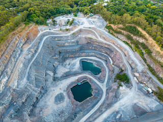 Aerial drone perspective of a vast open-cut quarry with deep blue-green water pits amidst a dense green bushland landscape. © Mujie