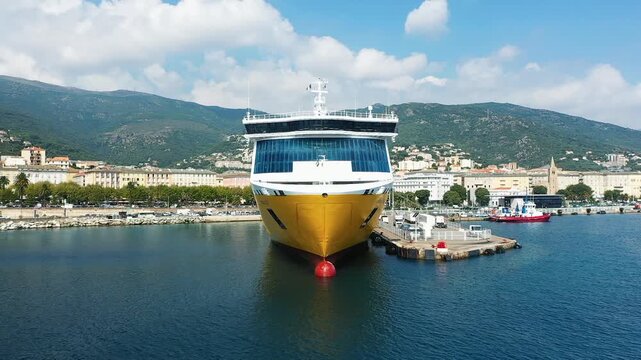 Large yellow ferry moored at the harbor in Bastia, Corsica, with mountains and city buildings in the background on a sunny day.