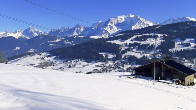 Scenic winter view of snowy fields, rustic barn, and the majestic Massif du Mont Blanc mountains under a clear blue sky. Idyllic alpine setting with traditional wooden chalet and distant village.