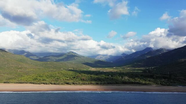 Aerial view of the Delta du Fango with sandy beach, lush greenery, and dramatic mountains under a partly cloudy sky in Corsica, France.