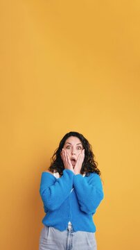 Expressive young woman standing with her mouth open in shock. Studio shot of a female model showing astonishment and disbelief on a solid yellow background. Vertical