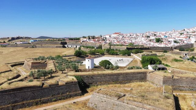 Aerial view of the historic fortifications and old town of Elvas, Portugal, featuring stone walls, a white tower, and a dry landscape under clear blue sky.