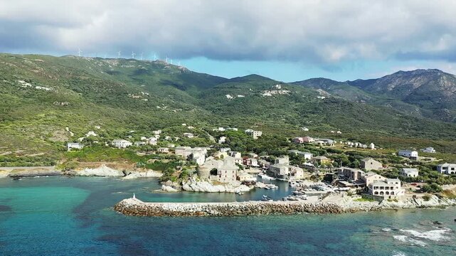 Scenic aerial view of the coastal village of Centuri in Corsica, France, with turquoise waters, a small harbor, and surrounding green hills under a cloudy sky.