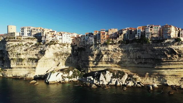 Dramatic limestone cliffs topped with historic buildings overlooking the Mediterranean Sea in Bonifacio, Corsica, under a clear blue sky.