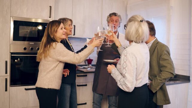 Group of cheerful senior friends gathered in a modern kitchen. They are smiling, talking, and making a toast with glasses of white wine