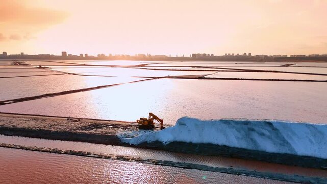 Sunset landscape with excavator working on salt production site. Golden hour view of industrial machinery and salt piles reflecting in water.