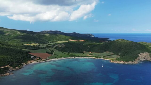 Scenic aerial view of Plage de Tamarone with turquoise waters, green hills, and farmland along the Corsican coastline under a partly cloudy sky.