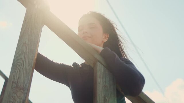 Young woman in casual outfit stands by wooden fence looking up in sunlight, calm confident lifestyle portrait. Nature, freedom, beauty, eco, lifestyle, sky, sunlight, calm, inspiration, femininity