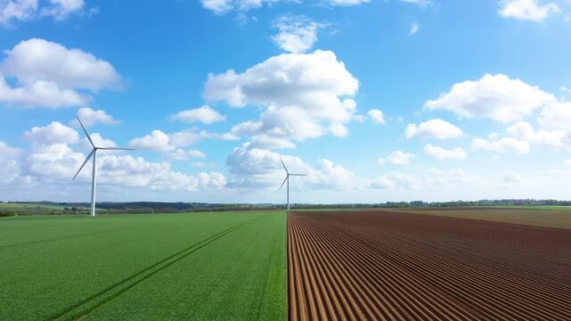 Aerial view of wind turbines standing between green and plowed fields under a blue sky with clouds in Normandy, France. Renewable energy and agriculture coexist in a rural landscape.