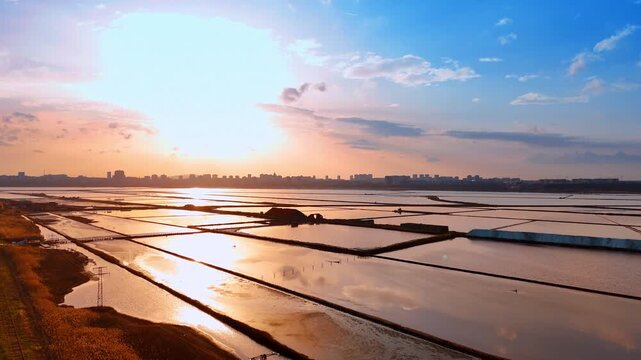 Aerial view of salt pans at sunset reflecting sky. Geometric ponds for salt production with sun reflection and city background.