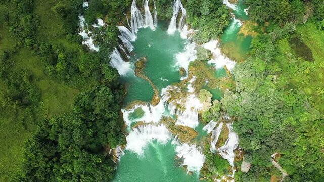 Stunning aerial shot of Ban Gioc Waterfall surrounded by lush green forest and turquoise water in Vietnam. The cascading falls create a breathtaking natural landscape.