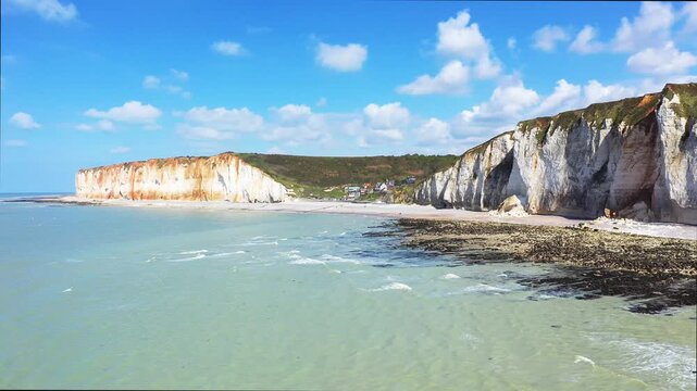 Scenic view of dramatic white chalk cliffs meeting the calm sea under a blue sky at Les Petites Dalles, Normandy, France. The coastline features rugged cliffs, small beach, and picturesque coastal