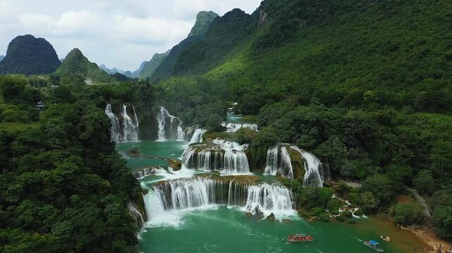 Scenic aerial view of Ban Gioc Waterfall cascading through lush green forest and mountains in northern Vietnam. Turquoise water flows over multiple tiers, creating a dramatic natural landscape.