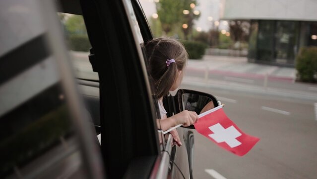 Little girl holding Switzerland flag, looking out the car window. Slow motion