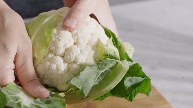 Hands holding fresh cauliflower with green leaves on a wooden cutting board. High quality 4k footage